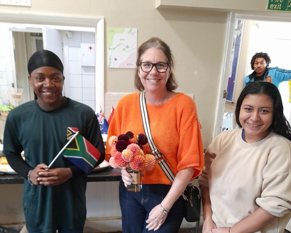 Three people looking at camera and smiling. The boy on the left is holding a Jamaican flag. The woman in the middle is holding a vase of dahlias