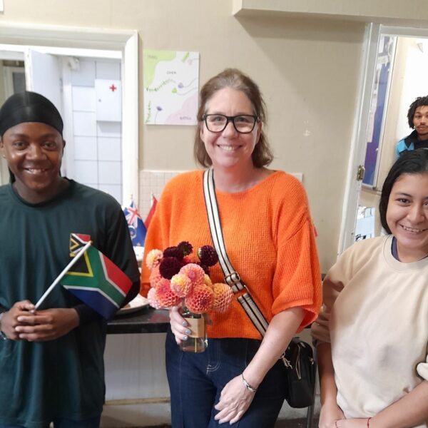 Three people looking at camera and smiling. The boy on the left is holding a Jamaican flag. The woman in the middle is holding a vase of dahlias