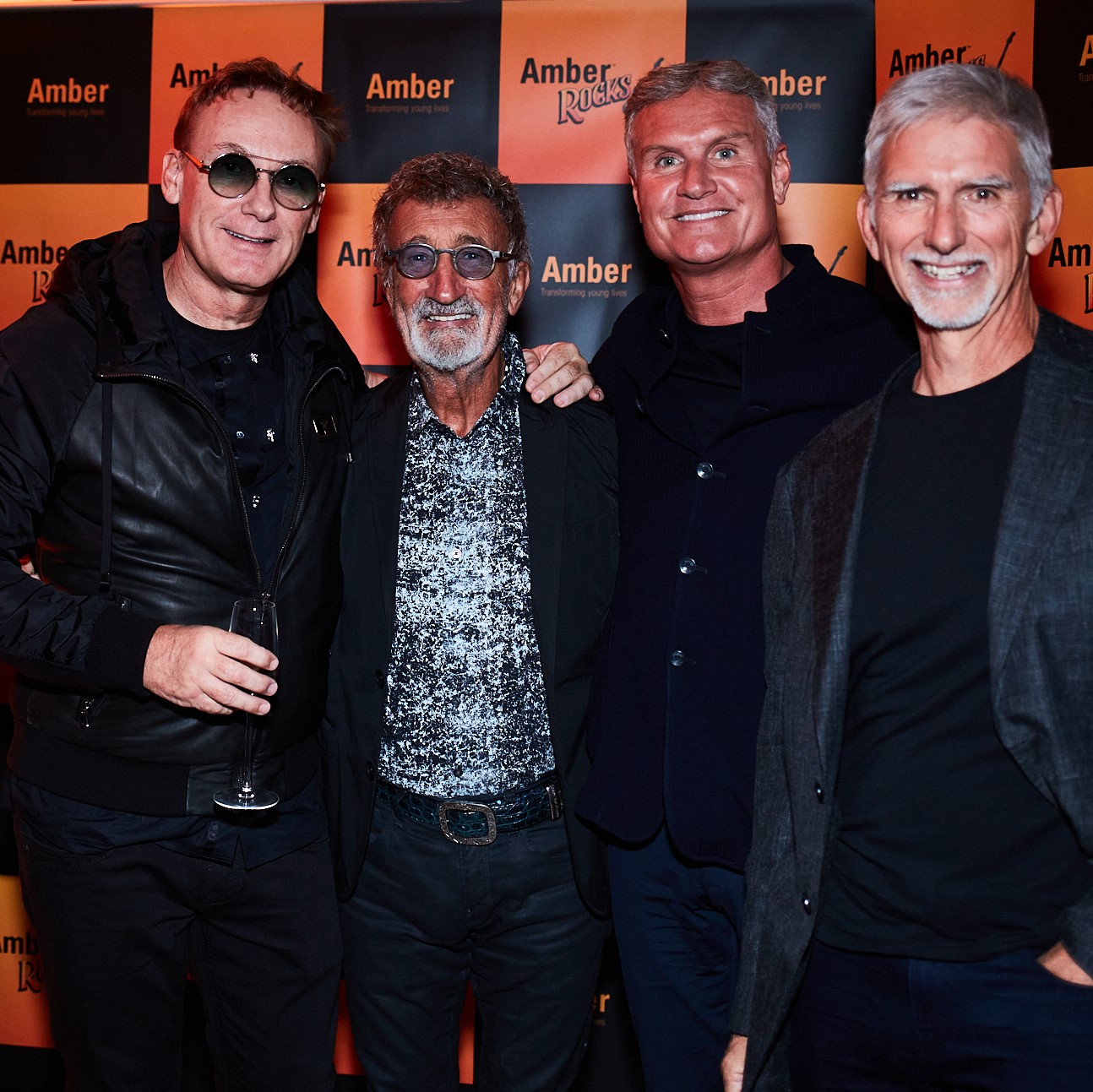 From left to right Bertrand Garchot, Eddie Jordan, David Coulthard and Damon Hill. All smiling at camera with an prange and black Amber Rocks background.